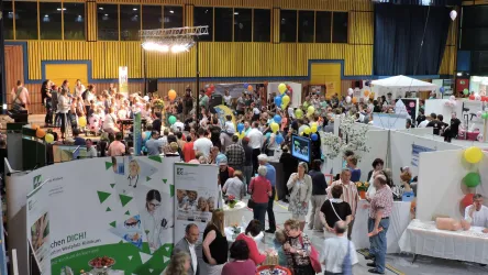 Eine lebhafte Messe mit vielen Besuchern und Ständen. Bunte Luftballons und Tische mit verschiedenen Auslagen schaffen eine festliche Atmosphäre.