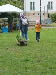Eine Person zieht einen kleinen Wagen mit Pflanzen, während ein Kind daneben geht. Im Hintergrund ist ein Marktstand unter einem Zelt zu sehen.