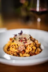 Ein köstlicher Teller Pasta mit buntem Gemüse und einer feinen Käseschicht obendrauf. Im Hintergrund ist ein Glas Rotwein zu sehen.