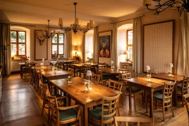 Ein gemütlicher Gastraum mit Holztischen und Stühlen. Fenster lassen warmes Licht herein und schaffen eine einladende Atmosphäre.