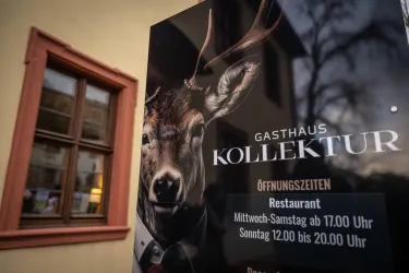 Ein Schild des Gasthauses Kollektur zeigt ein Reh mit Geweih. Daneben sind die Öffnungszeiten des Restaurants aufgelistet.