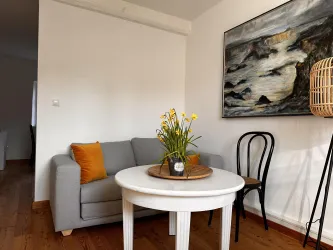 Ein gemütliches Wohnzimmer mit einem grauen Sofa und orangefarbenen Kissen. Auf dem Tisch steht eine Blumenvase, die den Raum lebendig macht.