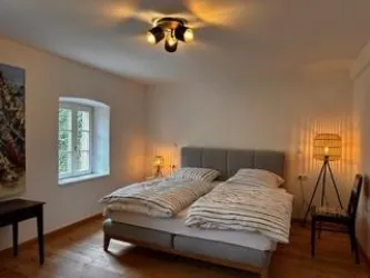 Ein gemütliches Schlafzimmer mit einem Doppelbett und sanftem Licht. Die Wände sind hell und ein Fenster sorgt für Tageslicht.