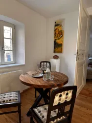 Ein gemütlicher Essbereich mit einem runden Holztisch und zwei Stühlen. An der Wand hängt ein farbenfrohes Bild, und durch das Fenster fällt natürliches Licht.