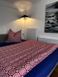 Ein gemütliches Schlafzimmer mit einem Bett und bunten Kissen. Neben dem Bett steht eine Lampe und an der Wand hängt ein modernes Bild.
