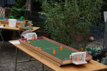 Ein kleines Tischfußballspiel mit orangefarbenen Figuren auf einem grünen Spielfeld. Im Hintergrund sind weitere Spielmaterialien und Pflanzen zu sehen.