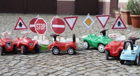 Eine Reihe von Spielzeugautos steht vor verschiedenen Verkehrszeichen auf einem Pflasterboden. Die Autos sind bunt und verkörpern eine kindgerechte Verkehrserziehung.