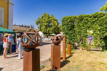 Ein Kunstmarkt mit Skulpturen aus Metall in einem begrünten Garten. Menschen genießen das sonnige Wetter und die Atmosphäre.