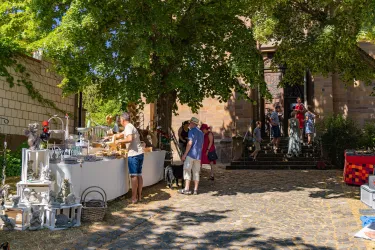 Ein lebhafter Markt unter einem großen Baum, wo Menschen Waren kaufen und verkaufen. Im Hintergrund ist ein historisches Gebäude sichtbar.