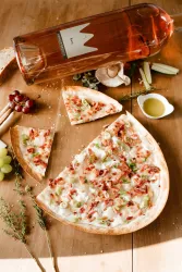 Eine leckere Pizza mit Schinken, Zwiebeln und frischen Kräutern liegt auf einem Holztablett. Daneben stehen ein Glas Wein und verschiedene frische Zutaten.