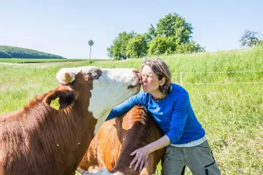 Eine Frau küsst eine Kuh auf einer grünen Wiese. Im Hintergrund sind Bäume und ein blauer Himmel sichtbar.