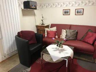 Ein gemütliches Wohnzimmer mit einem roten Sofa und einem schwarzen Sessel. Auf dem Tisch liegt eine weiße Decke mit einer Blumenvase.