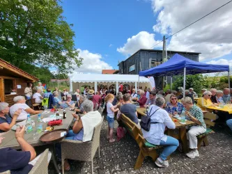 Eine große Gruppe von Menschen sitzt gemütlich im Freien, umgeben von Tischen und Bänken. Es ist ein sonniger Tag mit einigen Wolken und einer freundlichen Atmosphäre.