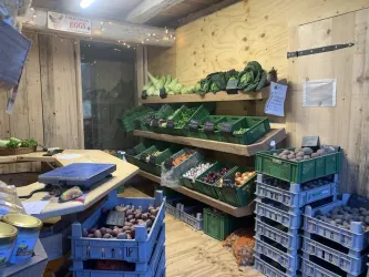 Ein kleiner Verkaufsstand mit frischem Gemüse und Obst. Regale sind mit verschiedenen Sorten gefüllt, während ein Tisch bereit zum Abwiegen von Produkten steht.