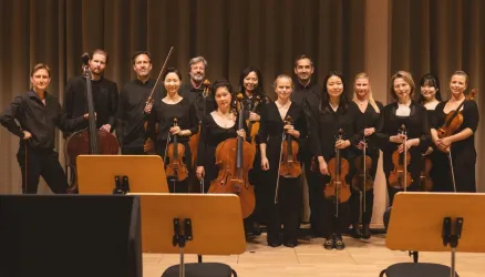 Eine Gruppe von Musikern steht auf der Bühne und hält Instrumente. Alle tragen schwarze Kleidung und lächeln in die Kamera.