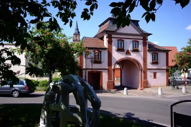 Ein historisches Gebäude mit einem charakteristischen Tor in einer kleinen Stadt. Im Vordergrund sind Bäume und eine Skulptur sichtbar.