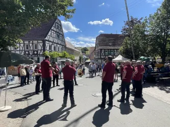 Eine Gruppe von Männern in roten Hemden steht auf der Straße, während sie einer Darbietung zusehen. Im Hintergrund gibt es ein traditionelles Fachwerkhaus und Bäume.