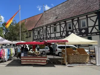 Ein lebhafter Markt mit mehreren Ständen und Besuchern. Im Hintergrund sind grüne Bäume und ein blauem Himmel zu sehen.
