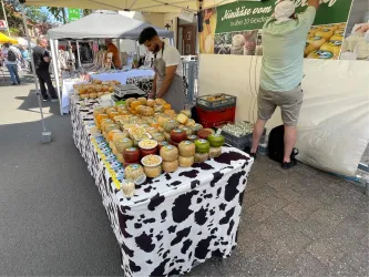 Ein Marktstand mit einem weißen Pavillon und verschiedenen handwerklichen Produkten. Im Hintergrund sind Menschen und ein schöner blauer Himmel zu sehen.