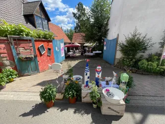 Eine gemütliche Gasse mit Blumendekorationen und Tischen im Freien. Im Hintergrund sind Menschen und bunte Sonnenschirme zu sehen.