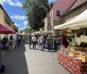 Ein belebter Marktplatz mit Ständen, die Taschen und Accessoires verkaufen. Im Hintergrund sieht man ein traditionelles Fachwerkhaus und blauem Himmel.