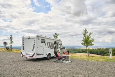 Ein Campingmobil steht auf einem ruhigen Platz mit Blick auf die Landschaft. Eine Person sitzt entspannt am Tisch und genießt die Natur.