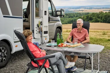Ein älteres Paar sitzt entspannt am Tisch vor ihrem Wohnmobil. Sie genießen eine Mahlzeit inmitten einer schönen Landschaft mit Feldern im Hintergrund.