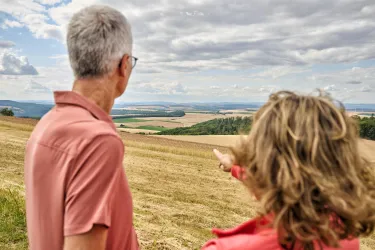 Ein Paar steht auf einem Hügel und blickt auf eine weite Landschaft. Der Himmel ist bewölkt und die Felder erstrecken sich bis zum Horizont.