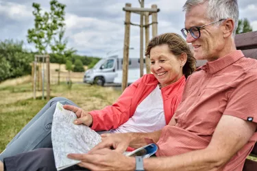 Ein Paar sitzt gemütlich auf einer Bank und schaut auf eine Landkarte. Im Hintergrund ist ein Wohnmobil und eine grüne Landschaft zu sehen.