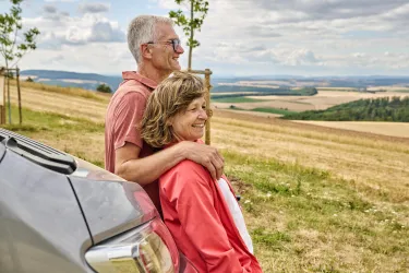 Ein glückliches Paar steht an einem Auto und schaut in die Ferne. Die Landschaft ist hügelig und wirkt ruhig und friedlich.