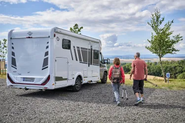 Ein Paar geht mit Stativ zu einem Wohnmobil auf einem Campingplatz. Im Hintergrund ist eine grüne Landschaft und ein blauer Himmel zu sehen.