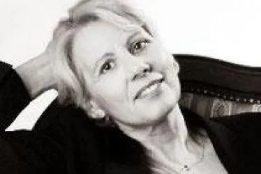 Eine Frau mit blonden Haaren sitzt entspannt auf einem Stuhl. Sie lächelt freundlich in die Kamera.