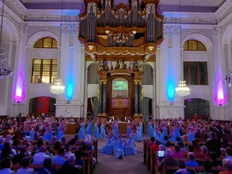 Eine festliche Aufführung in einer prächtigen Kirche mit einem großen Orgelretabel. Tänzerinnen in blauen Kleidern führen vor einem begeisterten Publikum auf.