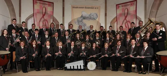Eine große Musikgruppe steht in formellen Anzügen zusammen. Im Hintergrund sind dekorative Banner zu sehen.