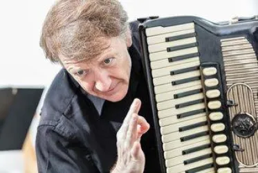 Ein Mann spielt Akkordeon und zeigt mit der Hand eine Geste. Er trägt ein schwarzes Hemd und hat lockiges Haar.