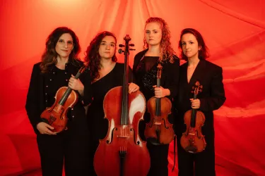 Vier Musikerinnen stehen vor einem roten Hintergrund und halten ihre Instrumente. Sie tragen elegante schwarze Kleidung und scheinen bereit für einen Auftritt zu sein.