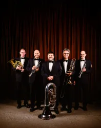 Eine Musikgruppe aus fünf Männern in eleganten Anzügen steht vor einem braunen Hintergrund. Sie halten verschiedene Blechblasinstrumente, darunter eine Tuba und eine Trompete.
