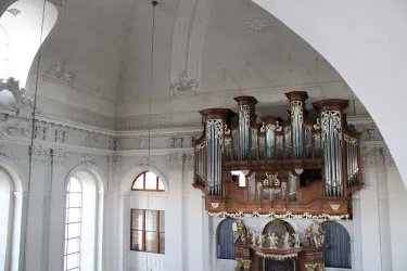 Eine beeindruckende Orgel in einer hellen Kirche mit kunstvoll verzierten Wänden. Die Fenster lassen viel Licht hinein und schaffen eine ruhige Atmosphäre.