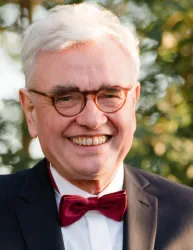 Ein lächelnder Mann mit grauen Haaren trägt einen schwarzen Anzug und eine rote Fliege. Im Hintergrund sind verschwommene grüne Bäume zu sehen.