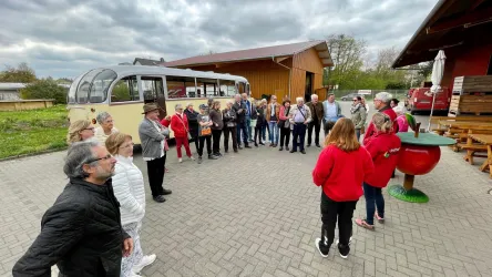 Eine Gruppe von Menschen steht in einem Kreis, während sie einem Vortrag hören. Im Hintergrund ist ein alter Bus und ein Holzgebäude zu sehen.