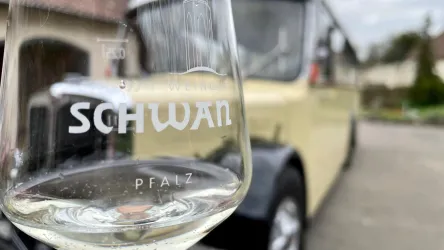 Ein Weinglas mit dem Aufdruck „SCHWAN PFALZ“ im Vordergrund. Im Hintergrund ist ein gelber Oldtimer-Bus zu sehen.