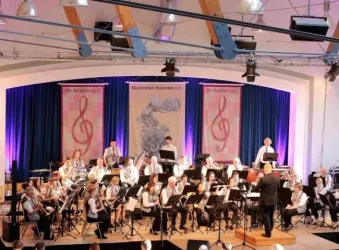 Ein Orchester spielt auf einer Bühne. Im Hintergrund sind dekorative Elemente mit Musiknoten und Lichtern zu sehen.