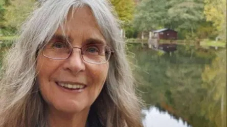 Eine lächelnde Frau mit grauem Haar steht vor einem ruhigen Gewässer. Im Hintergrund ist ein Holzhaus zwischen Bäumen zu sehen.