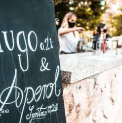 Ein schwarzes Schild mit der Aufschrift "Hugo 0,2 l & Aperol" steht an einer Bar. Im Hintergrund sieht man eine Person, die Getränke zubereitet.