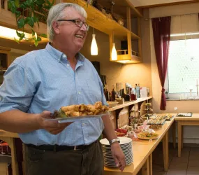 Ein lächelnder Mann hält einen Teller mit Essen in einer Einrichtung. Im Hintergrund ist ein Tisch mit verschiedenen Speisen und Getränken sichtbar.