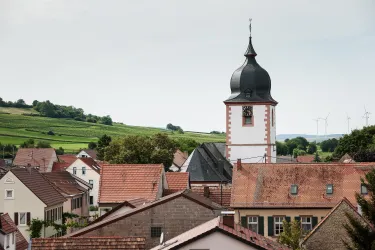 Eine malerische Dorfansicht mit einer markanten Kirchturm. Im Hintergrund sind sanfte Hügel und Windkraftanlagen zu sehen.