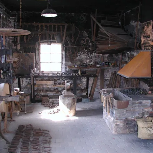 Eine alte Schmiede mit verschiedenen Werkzeugen und Maschinen. Der Raum wirkt rustikal und abgenutzt, mit einem offenen Fenster, das Tageslicht hereinflutet.