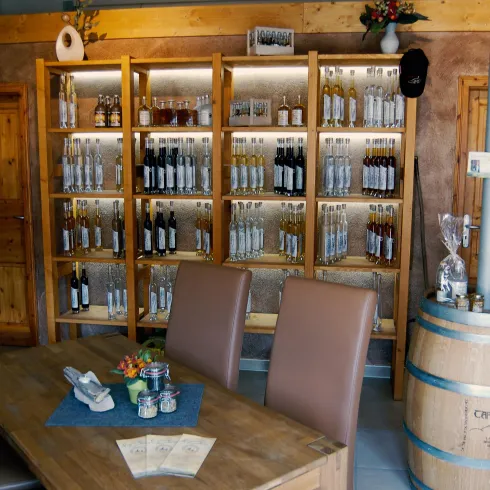 Ein gemütlicher Innenraum mit einem Holztisch und gepolsterten Stühlen. Im Hintergrund befindet sich ein Regal mit verschiedenen Getränken.