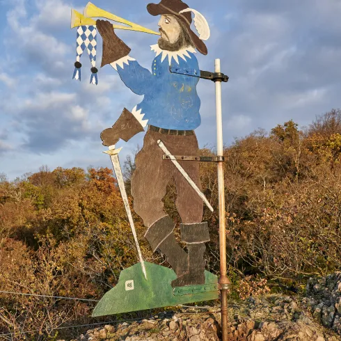 Eine bunte Holzskulptur eines Mannes, der ein Horn bläst. Im Hintergrund sind hügelige Landschaften und ein blauer Himmel zu sehen.