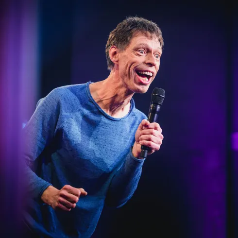 Ein Comedian steht auf der Bühne und spricht ins Mikrofon. Er lächelt und hat eine lebhafte Ausstrahlung.